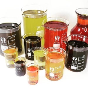 May include: Assortment of glass measuring cups filled with different colored liquids. The liquids are clear, yellow, orange, red, and black. The cups are marked with measurements in milliliters.
