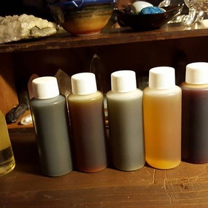 May include: Seven small bottles of essential oil, lined up on a wooden surface. The bottles are clear glass with white caps, except for the last bottle, which has a black cap. The oils are various colors, including clear, yellow, brown, and red.