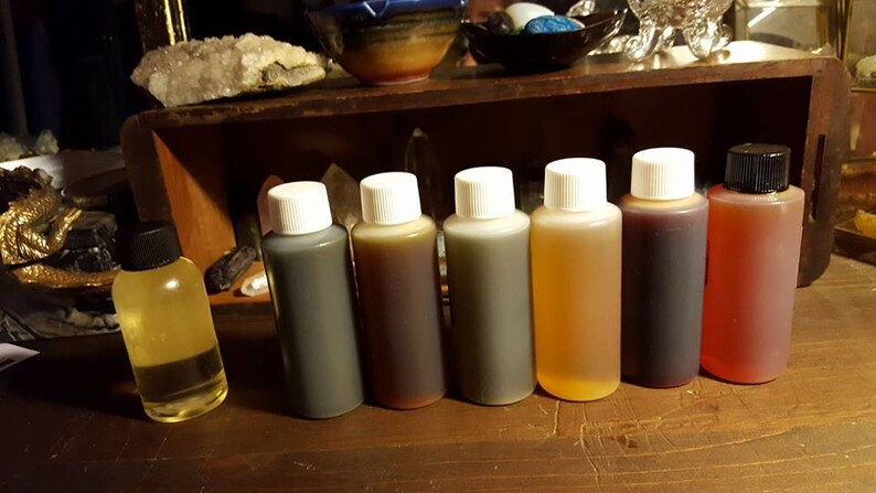 May include: Seven small bottles of essential oil, lined up on a wooden surface. The bottles are clear glass with white caps, except for the last bottle, which has a black cap. The oils are various colors, including clear, yellow, brown, and red.