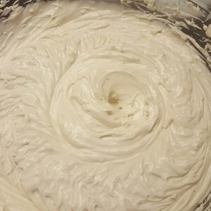 May include: A close-up of a white whipped cream frosting in a glass bowl. The frosting is smooth and creamy, with a swirl pattern.