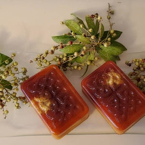 Puede incluir: Dos pastillas de jab&oacute;n rectangulares de color &aacute;mbar transl&uacute;cido con un patr&oacute;n de nudo celta en relieve en la superficie. Cada pastilla tiene una inclusi&oacute;n texturizada de color marr&oacute;n dorado en la parte superior y se muestra junto a ramas de hojas verdes y bayas.