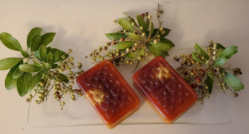 Puede incluir: Dos pastillas de jab&oacute;n rectangulares de color &aacute;mbar transl&uacute;cido con un patr&oacute;n de nudo celta en relieve en la superficie. Cada pastilla tiene una inclusi&oacute;n texturizada de color marr&oacute;n dorado en la parte superior y se muestra junto a ramas de hojas verdes y bayas.