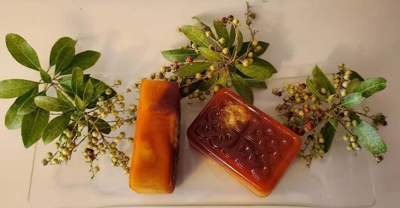 Puede incluir: Dos pastillas de jab&oacute;n rectangulares de color &aacute;mbar con un acabado transl&uacute;cido. Una pastilla presenta un dise&ntilde;o de nudo celta en relieve en su superficie. Las pastillas est&aacute;n dispuestas sobre una bandeja de cristal transparente y rodeadas de ramas con hojas verdes y peque&ntilde;as bayas.