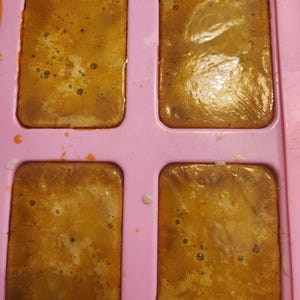 Puede incluir: Un molde de silicona rosa claro que contiene cuatro cavidades rectangulares llenas de una sustancia l&iacute;quida de color &aacute;mbar. La superficie del l&iacute;quido muestra peque&ntilde;as burbujas de aire y una textura ligeramente irregular y brillante.