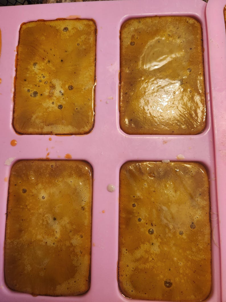 Puede incluir: Un molde de silicona rosa claro que contiene cuatro cavidades rectangulares llenas de una sustancia l&iacute;quida de color &aacute;mbar. La superficie del l&iacute;quido muestra peque&ntilde;as burbujas de aire y una textura ligeramente irregular y brillante.