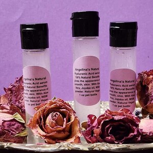 Puede incluir: Tres peque&ntilde;os frascos de vidrio transparente con tapas negras que contienen un suero de &aacute;cido hialur&oacute;nico. Los frascos est&aacute;n etiquetados con "Angelina's Natural Hyaluronic Acid Serum 100% Natural Benefits gives the appearance of smooth, skin. With Aloe Vera, Jojoba oil, MSM Powder. Natural Vegan 1 oz pet bottle". Los frascos est&aacute;n sobre una bandeja plateada con rosas rosadas secas.