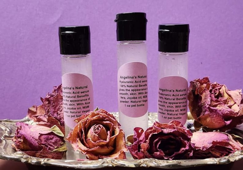 Puede incluir: Tres peque&ntilde;os frascos de vidrio transparente con tapas negras que contienen un suero de &aacute;cido hialur&oacute;nico. Los frascos est&aacute;n etiquetados con "Angelina's Natural Hyaluronic Acid Serum 100% Natural Benefits gives the appearance of smooth, skin. With Aloe Vera, Jojoba oil, MSM Powder. Natural Vegan 1 oz pet bottle". Los frascos est&aacute;n sobre una bandeja plateada con rosas rosadas secas.