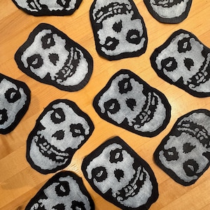May include: A collection of ten fabric patches, each featuring a silver-colored skull graphic printed on a black background. The skull designs are stylized with hollow eye sockets, a nose cavity, and a wide, grinning mouth with visible teeth.