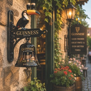 Può includere: Un'insegna da parete in metallo nero con un tucano che porta due bicchieri di birra sul becco, il testo GUINNESS ESTP 1759 e una campana nera appesa decorata con un'illustrazione dorata di un birrificio, il testo GUINNESS ST. JAMES'S GATE DUBLIN e un'arpa.