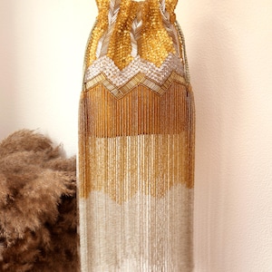 May include: A drawstring pouch bag featuring intricate gold and silver beadwork. The upper section displays a chevron pattern of gold and silver beads, while the lower portion consists of long, flowing gold and silver beaded fringe tassels.
