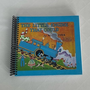 May include: A blue book with a spiral binding titled "The Little Engine That Could" by Watty Piper. The cover features a cartoon illustration of a blue train engine pulling a train up a hill. The book is a Platt & Munk Classic.