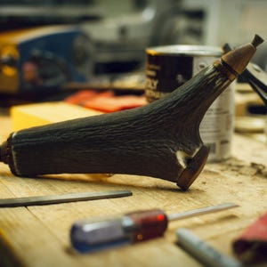 May include: A dark brown, textured antler powder flask featuring a metal nozzle at the tapered end and a circular metal loop at the base. The flask has a smooth, rounded wooden cap at the wider end, showcasing a natural, rugged, and organic surface.