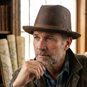 Puede incluir: Un sombrero fedora marrón con un acabado desgastado y envejecido, y una copa estructurada. El sombrero presenta una banda de color marrón oscuro alrededor de la base y un ala ligeramente curvada, hecho de un material liso con variaciones tonales.