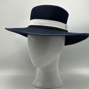 May include: A dark navy blue wide-brimmed hat featuring a structured crown and a flat brim. The hat is accented with a wide white ribbon band that sits above a thinner black ribbon band, encircling the base of the crown for a layered appearance.