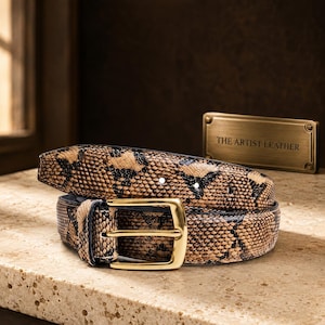 May include: A belt featuring a tan and black snakeskin pattern with a polished gold-tone metal buckle. A rectangular brass-coloured plaque positioned behind the belt displays the engraved text THE ARTIST LEATHER in black capital letters.