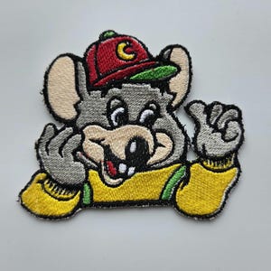 Puede incluir: Un parche bordado que muestra un personaje de ratón gris con una gorra de béisbol roja con un logotipo de media luna amarilla. El ratón tiene orejas grandes, nariz negra y viste una camisa amarilla de manga larga con detalles verdes.