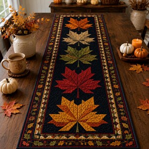 May include: A rectangular table runner featuring a black background with a central vertical row of five large, quilted maple leaves in shades of orange, beige, green, red, and yellow, bordered by a thin pattern of small autumn leaves.
