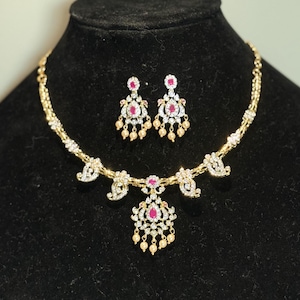 May include: A gold-toned necklace and matching earring set featuring teardrop-shaped pink stones, clear crystals, and small dangling cream-colored beads. The necklace has a central pendant and symmetrical paisley-inspired accents along the chain.