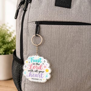 May include: A clear acrylic keychain with a scalloped edge attached to a grey backpack. The keychain features the colorful text Trust in the Lord with all your heart and PROVERBS 3:5, decorated with small stars, flowers, and swirl graphic accents.