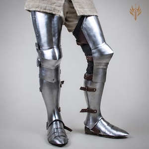 May include: Polished steel leg armor featuring articulated thigh, knee, and shin plates with brown leather straps and buckles. The set includes protective greaves and sabatons covering the feet, designed for full leg coverage and mobility.