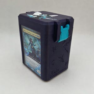 Puede incluir: Una caja de mazos rectangular de color morado oscuro con un cierre de color turquesa brillante. El frente tiene una ventana transparente que muestra una carta coleccionable titulada Aleta, Cunning Conqueror. La tapa tiene un gráfico de hada.