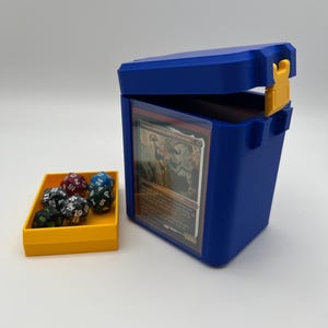 Puede incluir: Una caja de mazos de plástico azul con un cierre amarillo, que contiene una carta coleccionable visible a través de una ventana transparente. Junto a la caja hay una pequeña bandeja amarilla con seis dados de juego poliédricos de varios colores.