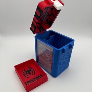 Puede incluir: Una caja para cartas de plástico azul y roja con tapa abatible que presenta un patrón de telaraña negra. Incluye una bandeja roja separada con un logotipo de araña negro y el texto SPIDER-MAN. Se ve una carta coleccionable en el interior.