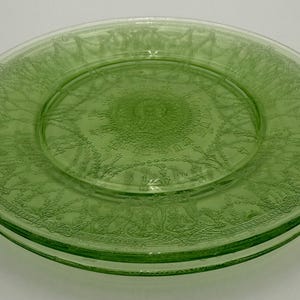 May include: A pair of stacked round light green glass plates featuring a detailed embossed pattern. The design includes a central sunburst motif surrounded by intricate garlands, swags, and beaded borders pressed into the translucent glass surface.