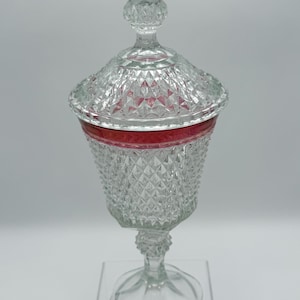 May include: A clear glass candy dish with a lid, featuring a diamond-cut pattern across the body and lid. The vessel includes a distinct horizontal band of deep red color near the rim and stands on a pedestal base with a pointed finial top.