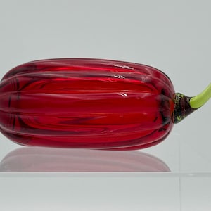 May include: A hand-blown glass sculpture shaped like a ribbed, elongated pumpkin. The main body is a deep, translucent red with subtle swirling patterns, while the stem is a smooth, bright lime green glass attached to one end of the piece.