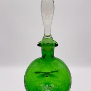 May include: A round, emerald green crackle glass decanter featuring a spherical body with a textured, web-like pattern and a clear, tapered glass stopper that fits into the narrow neck of the vessel, creating a distinct contrast in color and form.
