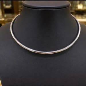 May include: A smooth, polished silver-toned metal collar necklace with a circular, rigid design. The necklace features a continuous, seamless band that rests in a rounded shape, showcasing a reflective, metallic surface without any visible clasps.