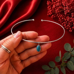 Op de afbeelding: Een zilverkleurige metalen choker-halsketting met een dunne, open cirkelvormige band en een enkele ovaalvormige turkoois hanger die aan een klein ringetje hangt. De hanger heeft een levendige blauwe kleur met donkerbruine matrix-aders.
