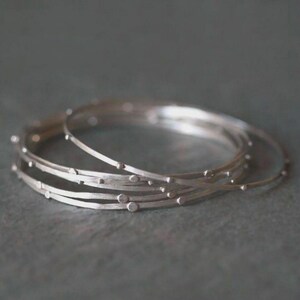 May include: A set of three thin, circular silver bangles with a matte finish. Each bangle features small, evenly spaced raised silver dots along the outer circumference, creating a textured appearance on the metallic surface of the jewelry.