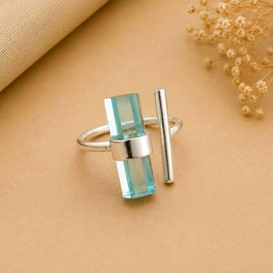 Puede incluir: Un anillo de plata con una piedra rectangular vertical de color azul claro translúcido engastada en una banda de metal pulido, acompañada de un elemento de barra de plata fino, paralelo y separado que se extiende.