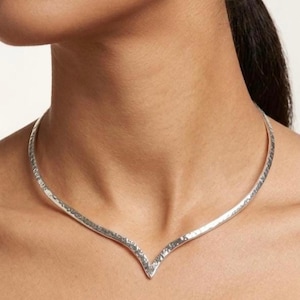 May include: A silver-toned V-shaped choker necklace featuring a textured, hammered surface finish. The rigid collar design sits close to the base of the neck, forming a distinct chevron point at the centre, crafted with a metallic sheen.
