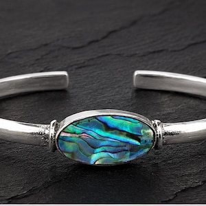 May include: A silver-toned cuff bracelet featuring a central oval-shaped inlay of iridescent abalone shell. The polished metal band has an open design, showcasing a smooth, reflective surface that contrasts with the vibrant blue and green colours.