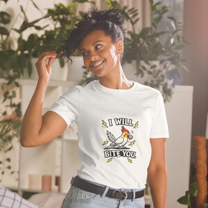 Peut inclure: Un t-shirt blanc à manches courtes avec un motif de calopsitte à crête rouge et joues jaunes. L'oiseau tient un petit objet dans son bec, entouré d'éclairs jaunes et du texte I WILL BITE YOU imprimé en lettres noires.