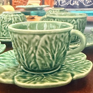 May include: A set of light green ceramic teacups and matching saucers, each featuring a detailed embossed leaf pattern that mimics the texture of cabbage leaves. The glazed finish gives the pieces a glossy appearance, with a rounded handle.