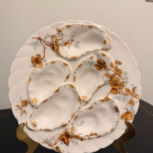 May include: A white porcelain oyster plate featuring five molded shell-shaped indentations. The plate is decorated with gold-painted edges and delicate floral patterns in shades of orange and light blue, with a scalloped rim design.