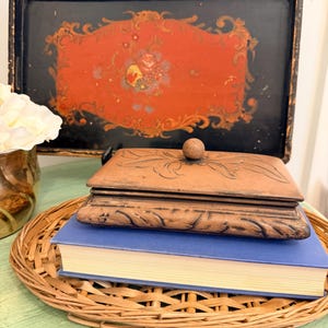 May include: A rectangular copper-toned metal box with a lid featuring a central knob and embossed leaf patterns, resting on a blue hardcover book. The book sits atop a woven wicker tray, positioned in front of a black and red painted metal tray.