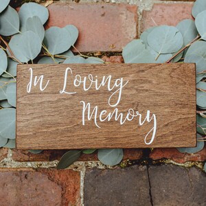 May include: A wooden sign with the text "In Loving Memory" written in white cursive lettering. The sign is surrounded by eucalyptus leaves.