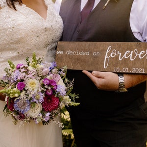 May include: A couple holds a wooden sign with the text "we decided on forever 10.01.2021". The bride is wearing a white lace dress and holding a bouquet of purple, white, and pink flowers.