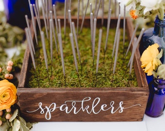 Op de afbeelding: Een houten doos gevuld met zilveren vuurwerk, met een bord met de tekst "sparklers". De doos staat op een wit oppervlak, omringd door gele bloemen en blauwe glazen flessen.
