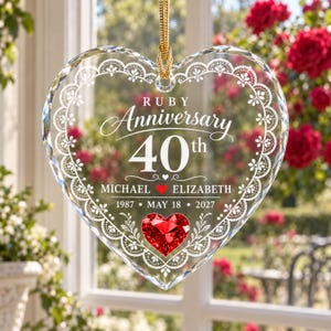 以下が含まれることがあります： 白いレース模様の縁取りと「Ruby Anniversary 40th」「Michael」「Elizabeth」「1987」「May 18」「2027」の文字が刻まれた透明なハート型のガラス製オーナメント。下部中央には赤いハート型のクリスタルが配置され、上部には金色の紐が付いています。