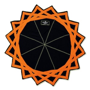 Op de afbeelding: Een cirkelvormig zwart kledingstuk met een levendige oranje geometrische stervormige overlay met puntige randen. Het midden toont een zwart label met de witte tekst "Flowchakra". Dunne, lichtgekleurde zigzagstiksels stralen vanuit het midden.