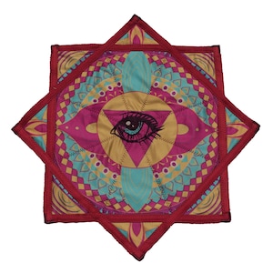 Può includere: Un sottobicchiere quadrato in tessuto trapuntato con una grafica centrale di un occhio circondato da un motivo geometrico a mandala nei toni del magenta, turchese e oro. I bordi sono rifiniti con una cucitura rosso scuro a stella.