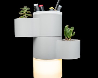 May include: A multi-tiered, light gray cylindrical organizer featuring three distinct sections for storage and planting. The top section holds pens, while the middle and upper side sections contain small green succulents. The base emits a warm white glow.