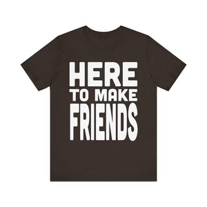 May include: A dark brown short-sleeved t-shirt featuring the white text "HERE TO MAKE FRIENDS" printed in a bold, sans-serif font across the center of the chest. The garment has a crew neckline and a standard fit with short sleeves.