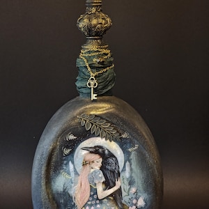Könnte beinhalten: Eine dekorative Glasflasche mit dunkler, strukturierter Oberfläche und einer bemalten Illustration einer Person mit langem rosa Haar, die einen großen schwarzen Raben unter einem Vollmond umarmt. Der Flaschenhals ist mit dunklem Stoff umwickelt.